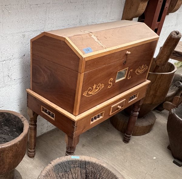 Mueble de madera con tapa inclinada, motivos claros en el frente y soporte de patas torneadas, junto a recipientes de madera.