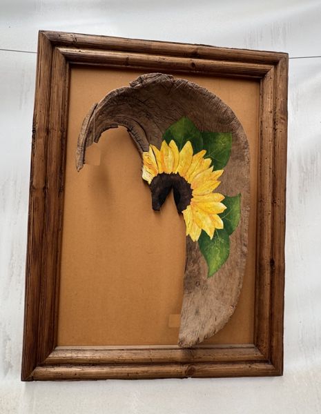 Marco de madera con fondo ocre y una pieza curva de madera pintada con un girasol amarillo y hojas verdes.
