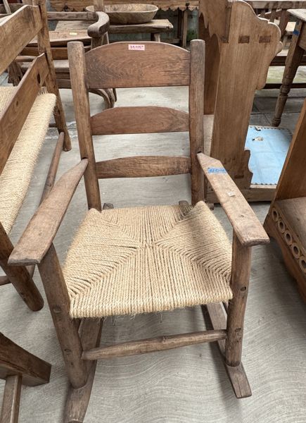 Mecedora de madera con respaldo de listones, brazos largos, asiento tejido claro y patines curvos; un patín alcanza el borde inferior.