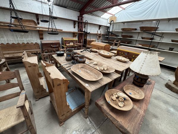 Interior con mesas y sillas de madera, recipientes y bandejas, una lámpara, estantes y estructura de cubierta visible.