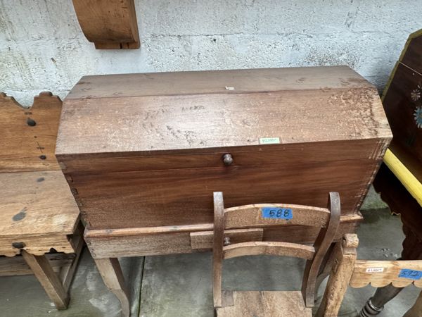Mueble marrón con superficie superior inclinada, pequeño herraje central y base saliente, parcialmente cubierto por una silla.