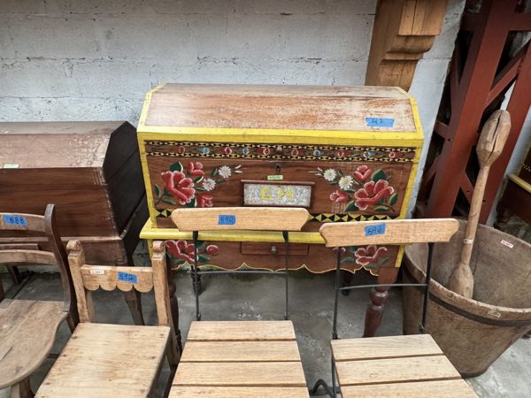 Mueble de frente oscuro con flores rojas, hojas verdes y bordes amarillos; varias sillas se superponen en primer plano.