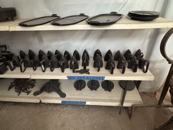 Estantes con piezas metálicas redondas en la parte superior, una hilera de planchas con asas al centro y pequeños herrajes debajo.