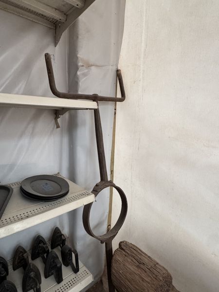 Estructura metálica estrecha junto a una estantería, con extremos superiores curvados hacia arriba, una barra vertical y un aro ovalado; la parte inferior queda fuera del encuadre.