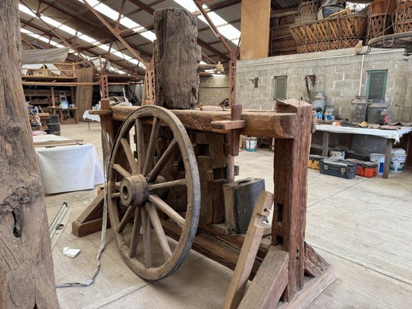 Gran rueda de madera con radios, aro metálico oscuro y cubo central, situada delante de una estructura de vigas; una correa clara pasa por la parte superior.