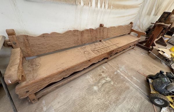 Banca larga de madera con respaldo y faldón de bordes ondulados, asiento de tabla y brazos; una tira de madera suelta descansa sobre el asiento.