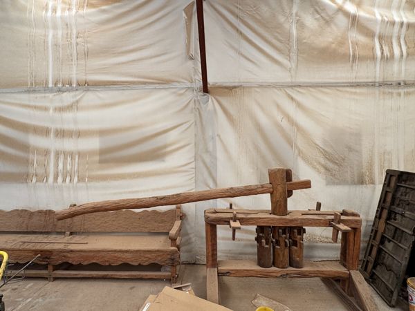 Estructura de madera con postes, travesaños, tres componentes cilíndricos verticales y una barra superior larga; parte de una banca aparece a la izquierda.