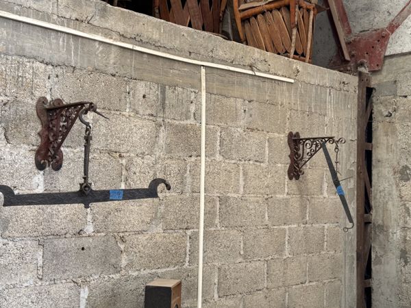 Dos soportes de pared con volutas y elementos metálicos suspendidos sobre un muro de bloques; el elemento izquierdo continúa fuera del encuadre.