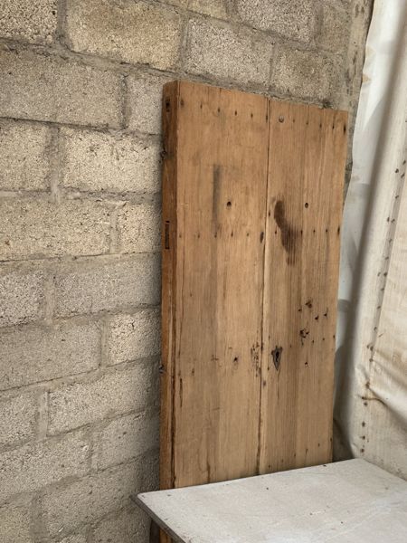 Panel de tablas verticales apoyado contra un muro de bloques junto a una tela blanca, con la parte inferior tapada por una superficie en primer plano.
