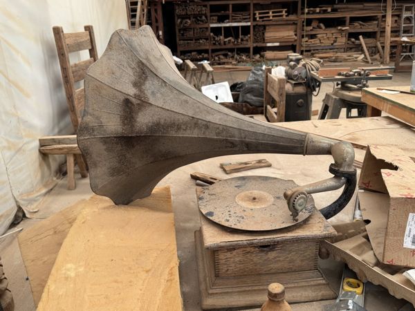 Gramófono con gran bocina metálica, tubo curvo, plato circular visible y caja de madera sobre una superficie con otros objetos.