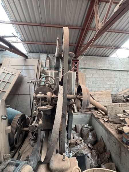 Vista oblicua parcial de maquinaria metálica con ruedas de radios, poleas ranuradas, cadena, ejes y bandas dentadas verticales entre materiales almacenados.