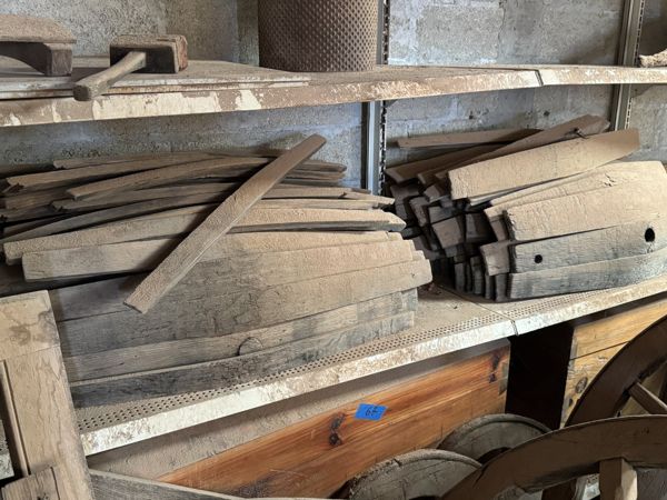 Tablas anchas, piezas gruesas con orificios o muescas y listones diagonales en una estantería metálica, con una caja de madera debajo y bastidores superpuestos.
