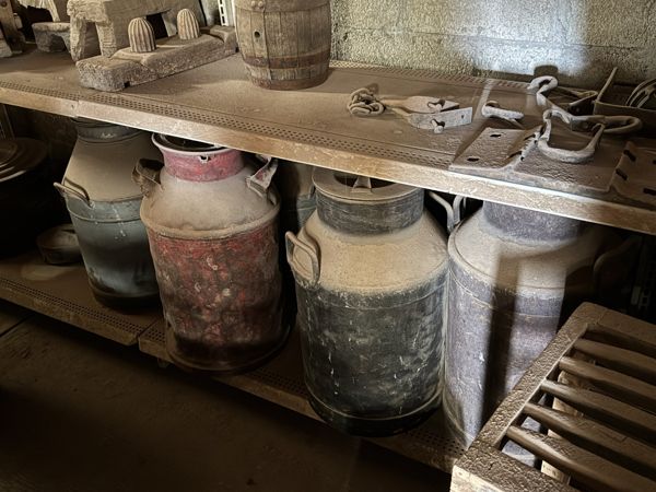 Varios recipientes metálicos tipo lechera, con hombros redondeados, cuellos estrechos y asas laterales, en una repisa inferior bajo piezas de madera y metal.