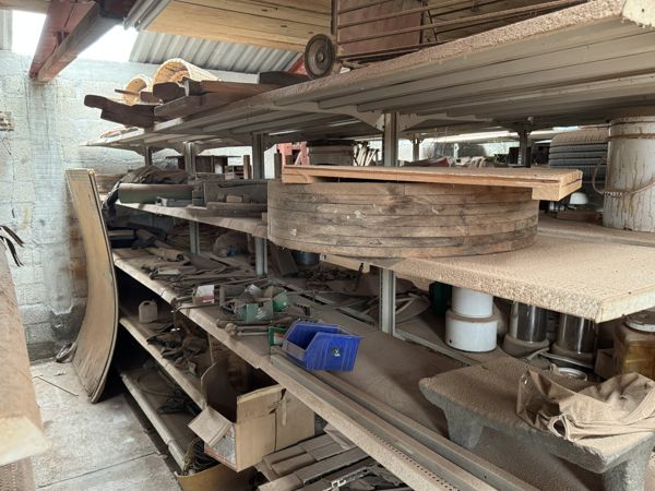 Gran forma circular de madera con juntas visibles y una pieza plana sobresaliente arriba, rodeada de tablas y objetos almacenados.