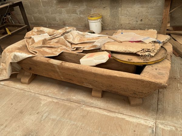 Contenedor bajo rectangular de madera sobre apoyos cortos, con la abertura superior cubierta parcialmente por telas y objetos apilados.