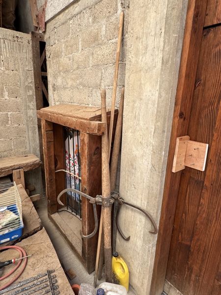 Mangos largos de madera con piezas metálicas curvas, junto a una estructura de madera, un muro de mampostería y una puerta de tablones.