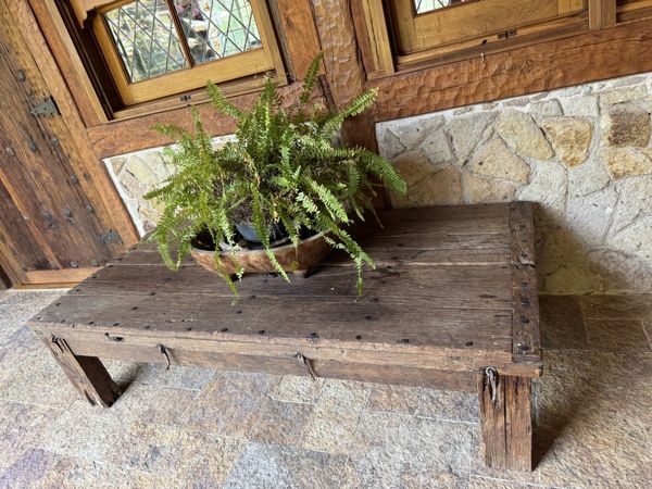 Mesa de madera de tablones gruesos con un helecho, junto a puertas de madera y un muro de piedra.