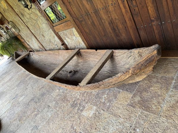 Vista oblicua del interior de una pieza alargada de madera con forma de canoa, bordes gruesos y tres travesaños, junto a una puerta de madera y piso de piedra.