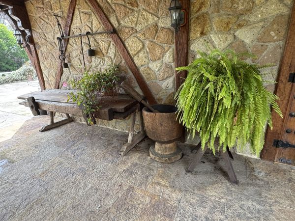 Estructura baja de madera, conjunto metálico colgante, recipiente de madera sobre pedestal y plantas en maceta frente a un muro de piedra con vigas y faroles.
