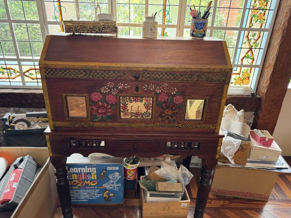 Mueble de madera con motivos florales pintados, bordes dorados, paneles rectangulares y patas torneadas, fotografiado frente a una ventana con cajas y objetos alrededor.