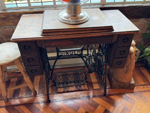 Mueble de madera con cajones laterales, bastidor metálico negro, pedal y rueda visibles, con la palabra Singer en el soporte y varios objetos sobre la cubierta.