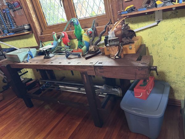 Banco de trabajo de madera con tornillo lateral y patas robustas, cubierto de figuras de aves y otros objetos, frente a ventanas y estantes de herramientas.